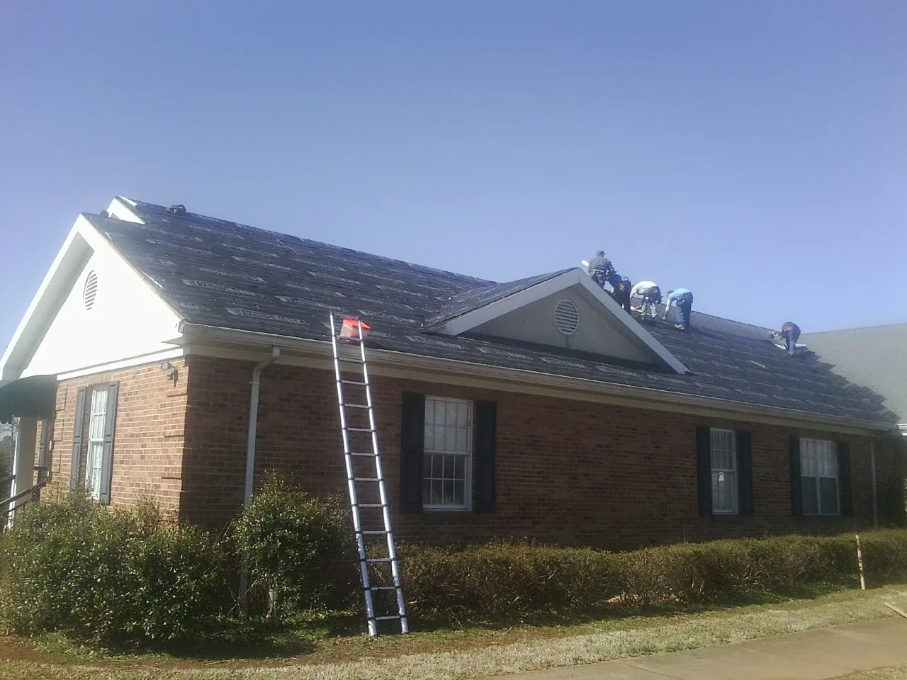 Crew tearing shingles off a brick ranch roof with a ladder set — Emergency Roof Repair work by our crew in Buies Creek