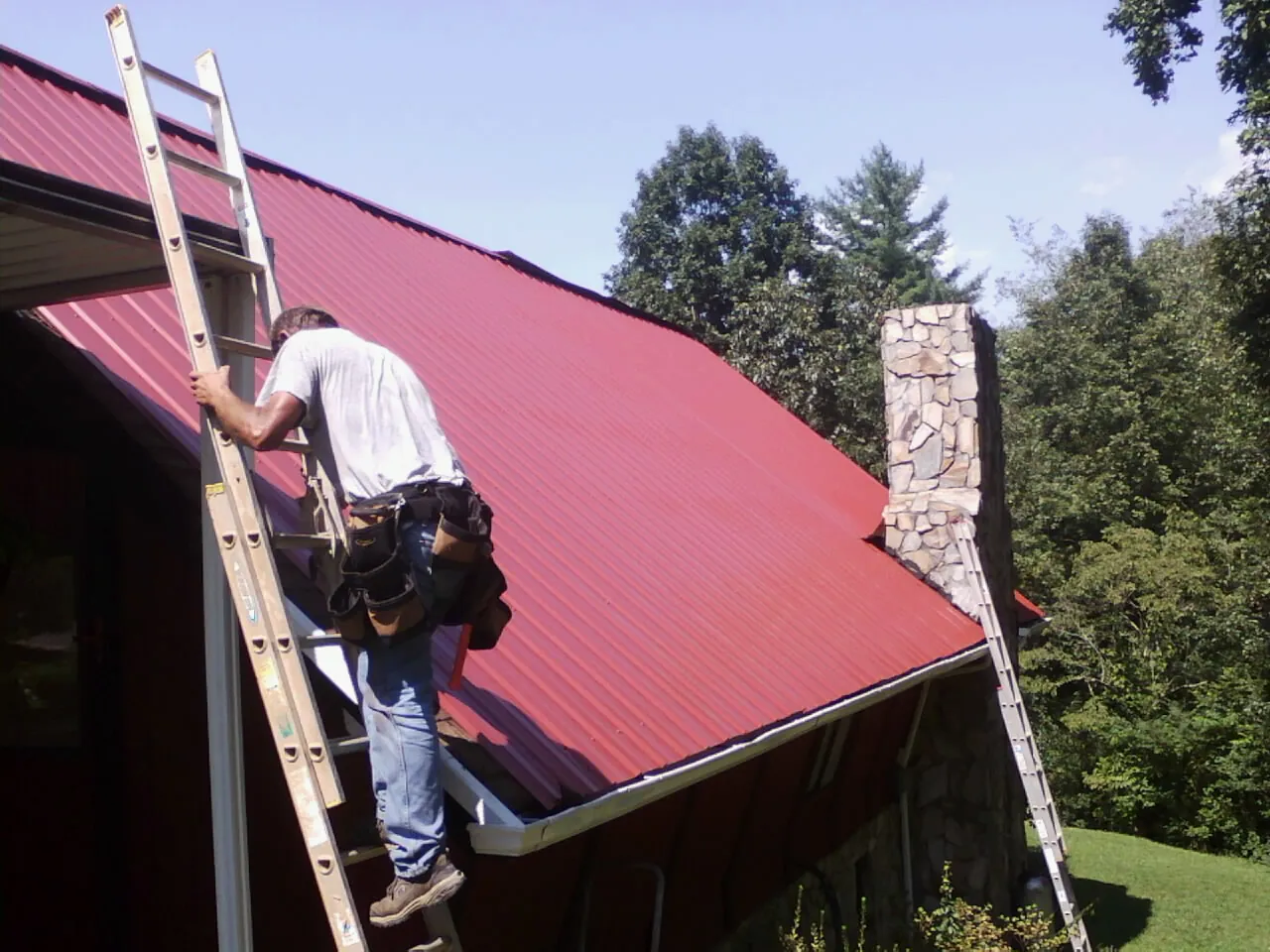 Roofer on a ladder beside a red metal roof and stone chimney — Soffit & Fascia Repair by Roofing Brothers in Buies Creek